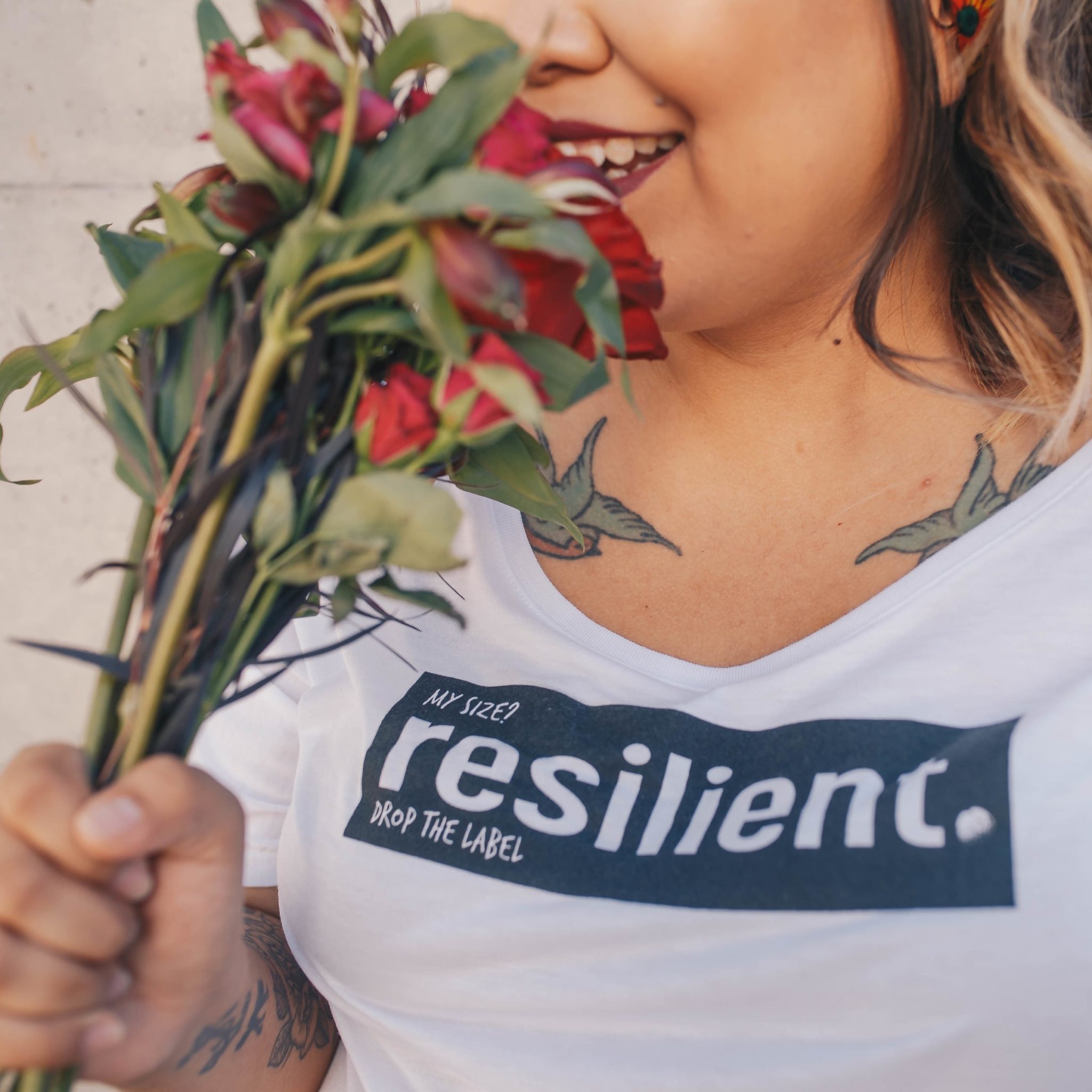 gruba dziewczyna, widoczna na zdjęciu od nosa do piersi, wącha czerwone kwiaty. Na obojczykach ma wytatuowane jaskółki, a na bluzce napis "my size? resilient.".