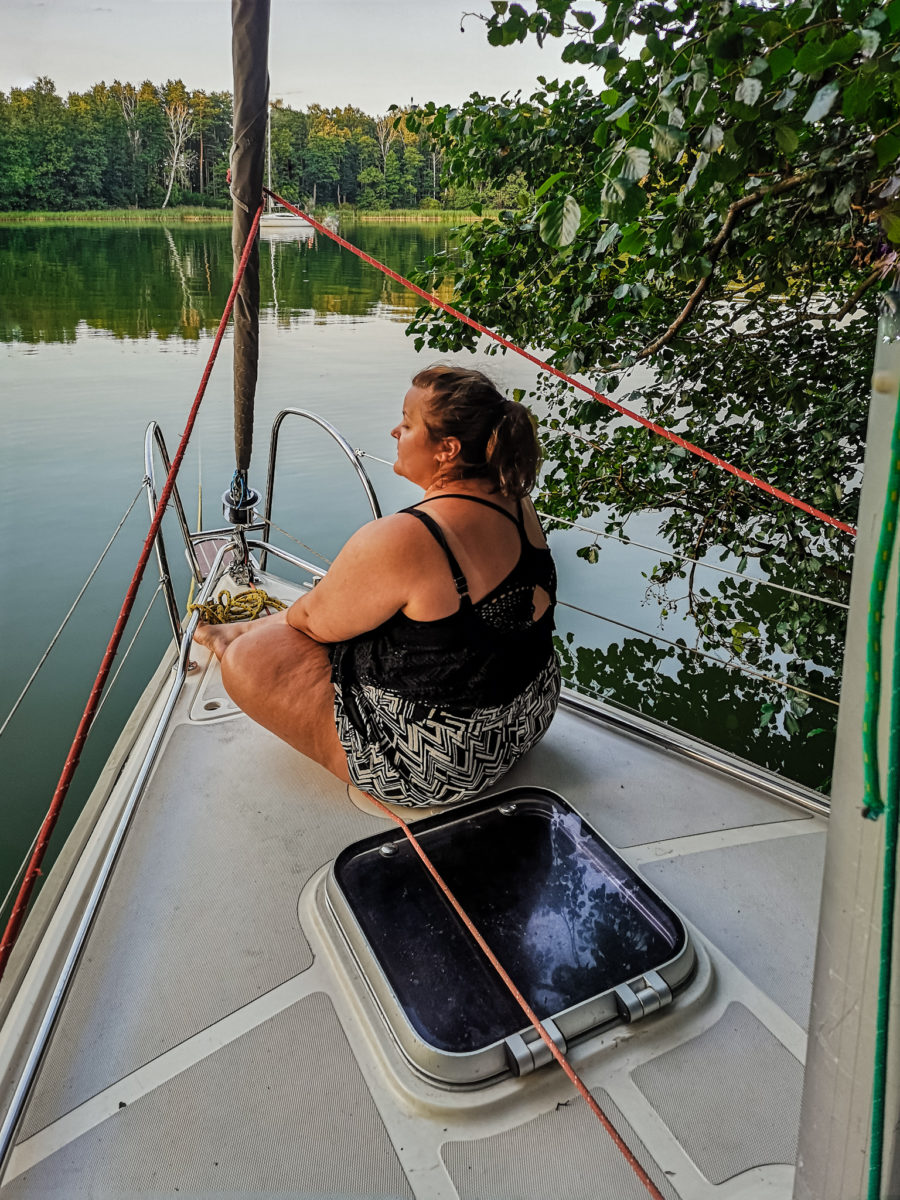 Ula siedząca na dziobie łódki, zwrócona tyłem do kamery, w tle mazurski krajobraz