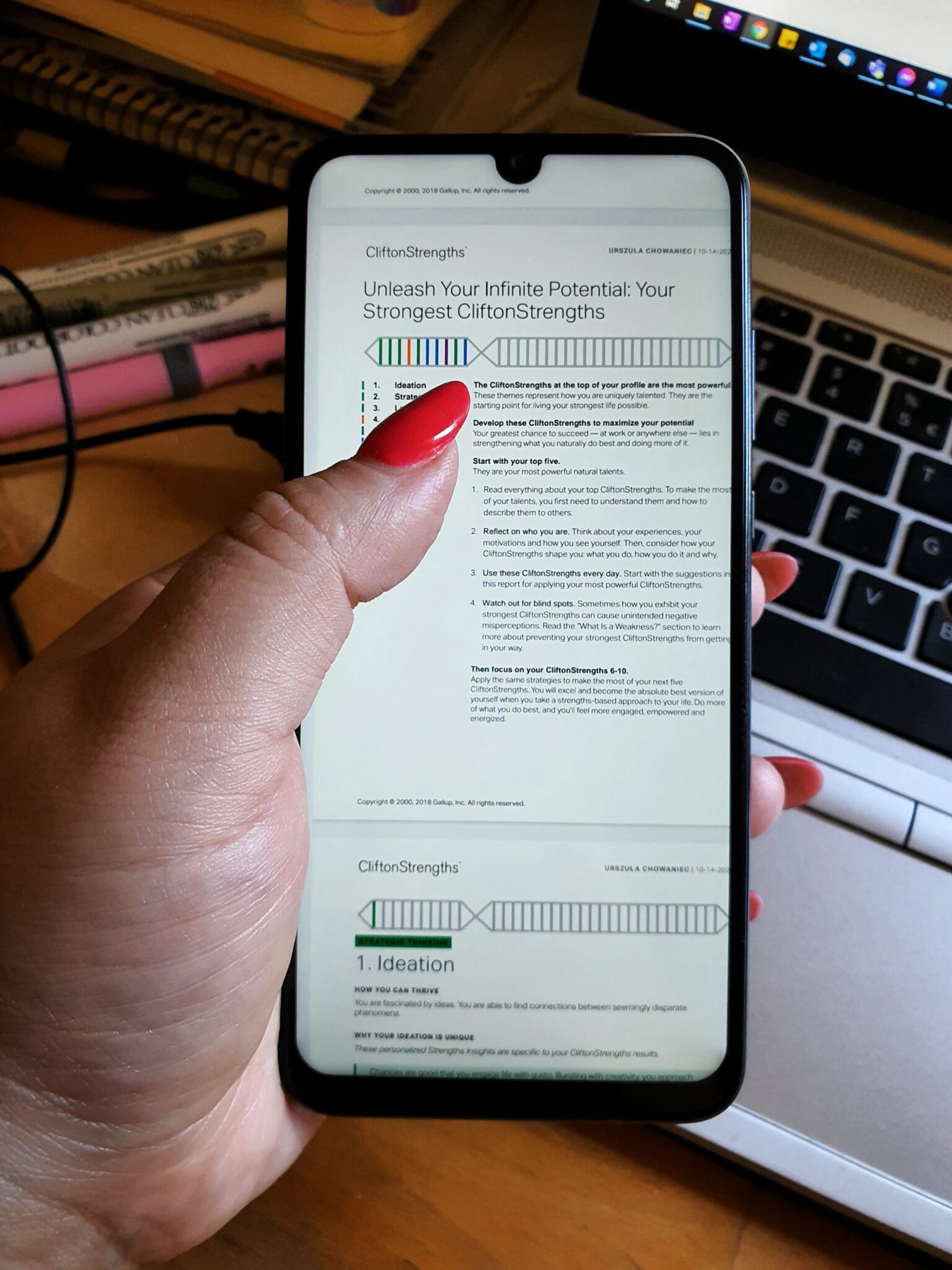 zdjęcie raportu z testu Gallupa widoczne na ekranie trzymanego przez Ulę smartfona, w tle klawiatura laptopa i bałagan na biurku