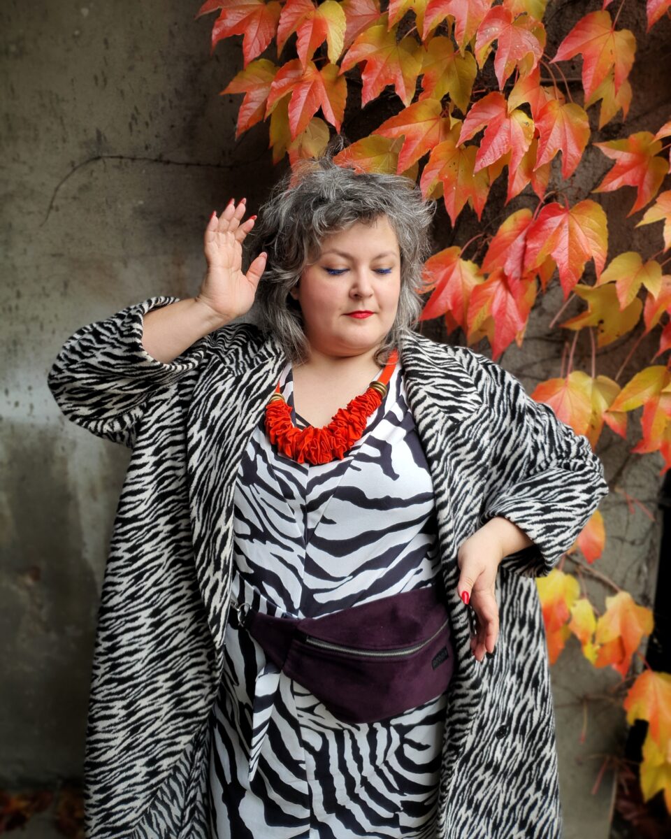 Ubrana w sukienkę i płaszcz Ula pozuje na tle betonowej ściany porośniętej jesiennym pnączem
