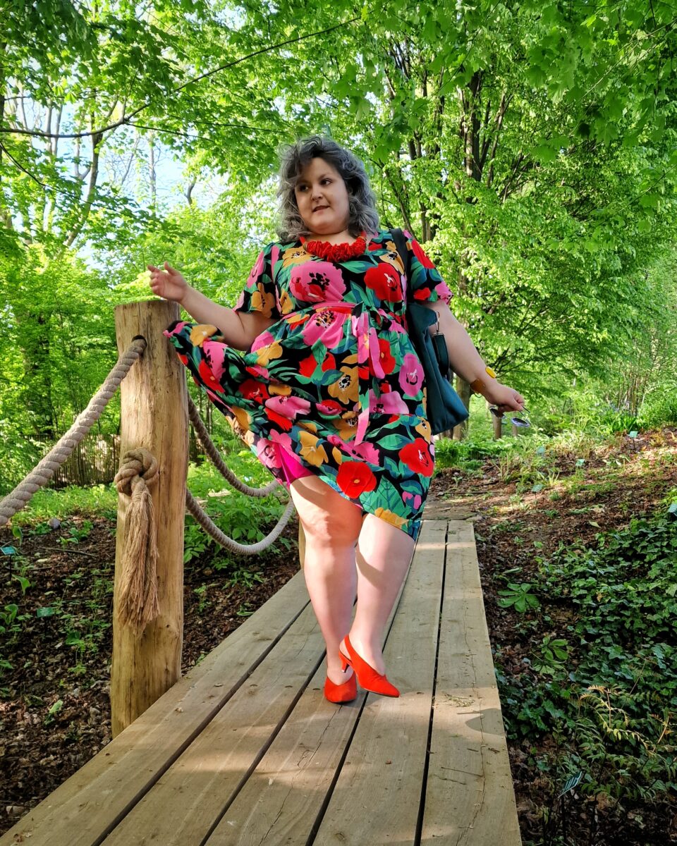 Dopamine dressing w wersji dla grubych - Ula w kolorowek sukience w kwiaty stoi na drewnianym mostku twarzą do aparatu, za nią widać dużo drzew i zieleni